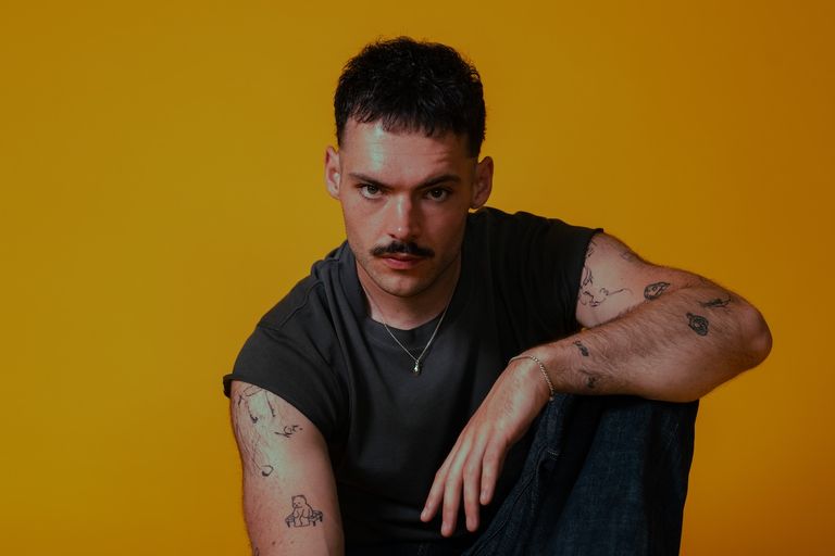 Studio portrait of musician James Marriott with arm tattoos, wearing a dark grey t-shirt against a vibrant yellow backdrop.